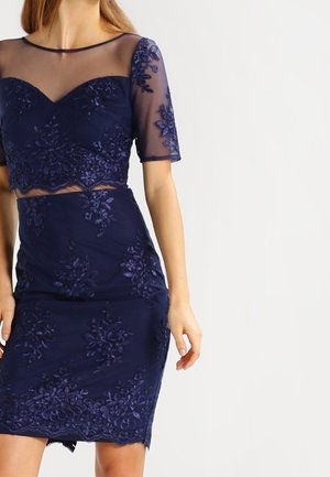 Frau trägt ein eng anliegendes marineblaues Spitzenkleid mit transparenten kurzen Ärmeln und floraler Stickerei, steht vor einem schlichten weißen Hintergrund.