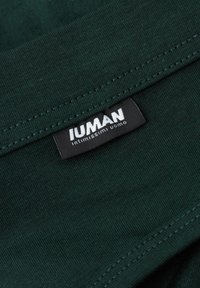 Tela verde oscuro con una textura suave y costuras visibles; presenta una etiqueta negra con texto blanco que dice "IUMAN intimi uomo."