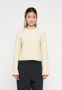 Cremefarbener gerippter Strickcardigan mit rundem Halsausschnitt, langen Ärmeln, Fronttaschen und Knopfverschluss. Getragen über dunkel gestreiften Hosen.