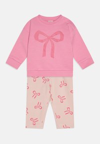 Rosa Sweatshirt mit einer großen Bogen-Grafik, kombiniert mit hellbeigen Leggings, die kleine rosa Bogenmuster aufweisen. Weicher Stoff, lässiges Design.