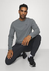 Szürke, hosszú ujjú póló, kerek nyakkivágással és fekete Nike logóval, sötét nadrággal és fekete sportcipővel párosítva, laza testtartást mutatva.