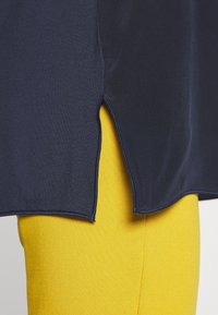 Granatowa bluzka z bocznymi rozcięciami, wykonana z gładkiego materiału, zestawiona z jasnżółtymi, teksturowanymi legginsami. Zbliżenie pokazujące szczegóły.