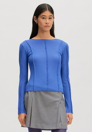 Woman with long dark hair wears a fitted blue long-sleeve top and a high-waist pleated black-and-white checkered skirt.