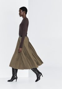 Femme marchant vêtue d'un haut à manches longues marron, d'une jupe midi olive et de bottes en cuir noires à talons hauts sur un fond blanc.