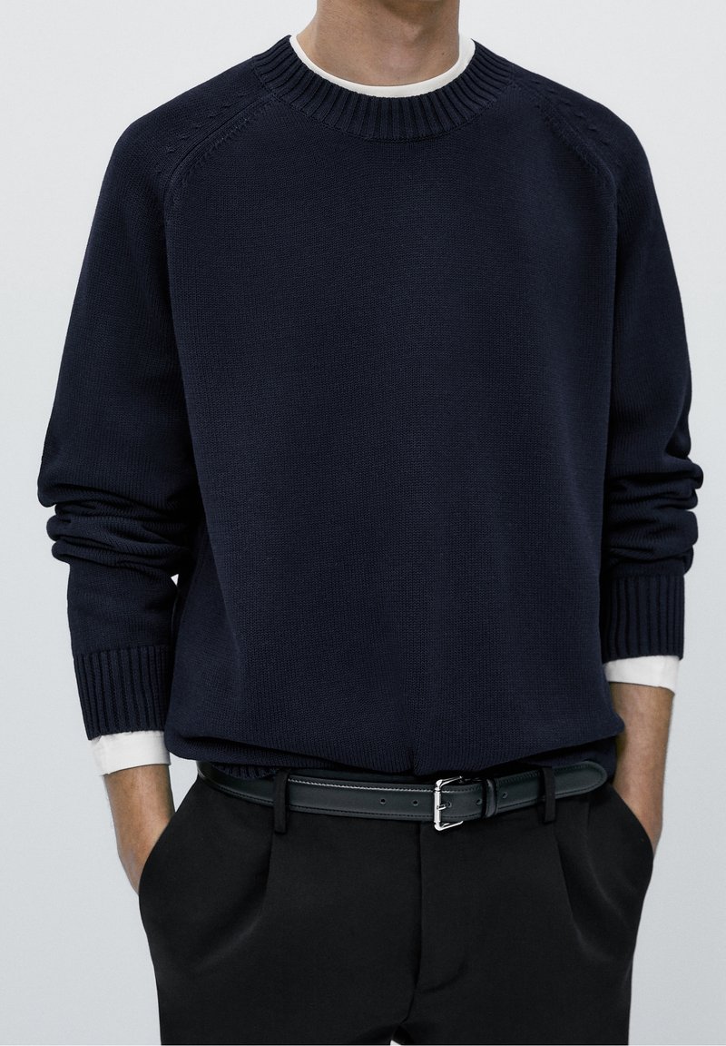Pull en maille bleu marine avec col et poignets côtelés. Porté sur une chemise à col blanc, associé à un pantalon foncé et une ceinture noire.