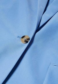 Blazer bleu clair, tissu lisse, bouton métallique unique, détail de couture subtil, revers aiguisés et poche avant.