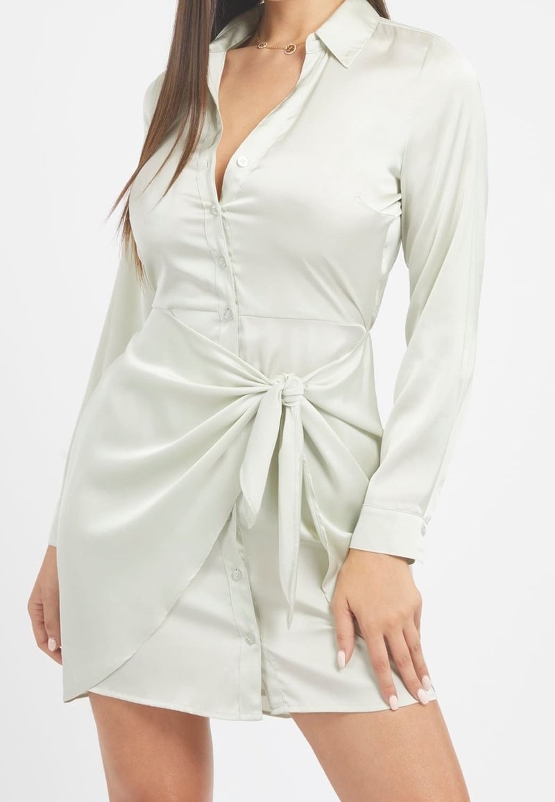 Nainen, joka on pukeutunut pitkähihamaiseen, vaaleasta satiinista valmistettuun paitamekkoon, jossa on napit ja sidottu vyötäröyksityiskohta, seisoo valkoista taustaa vasten.