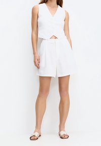 Weißes Leinen-Crop-Top mit V-Ausschnitt und vorne liegender Knopfleiste, kombiniert mit passenden locker sitzenden Shorts. Flache Sandalen vervollständigen das Outfit.