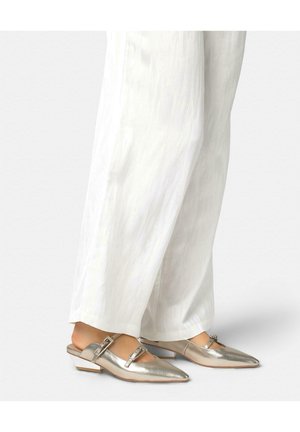 Pies con zapatos slingback plateados metálicos de punta puntiaguda con doble hebilla, combinados con pantalones blancos sueltos y texturizados.