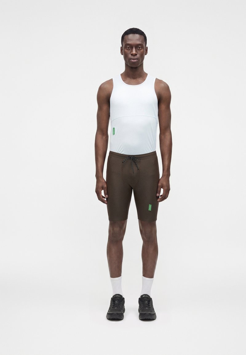 Homme debout face à l’avant, portant un haut blanc sans manches, un short marron à cordon, des chaussettes blanches et des baskets noires sur fond blanc.