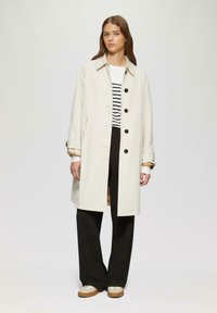 Heller Creme-Trenchcoat mit Kragen, Frontknöpfen und Klappentaschen, getragen über einem gestreiften Oberteil und schwarzen weitgeschnittenen Hosen.