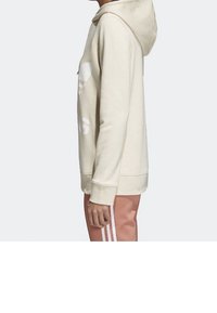 Béžová mikina s kapucí a volným střihem, která má na přední straně velké bílé logo, vyrobená z měkké látky, s žebrovanými manžetami a lemem.