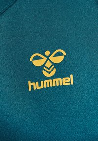 Gult stiliseret bi-logo og "hummel" tekst på teal tekstureret stof, sandsynligvis et sportstøj.