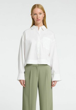 Camicia bianca cropped con bottoni, a maniche lunghe e una tasca sul petto, abbinata a pantaloni verdi oliva a vita alta. Tessuto morbido.