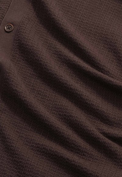 Brauner, strukturierter Strickstoff mit einem Knopf und geripptem Rand, der Falten und Schatten Details zeigt.