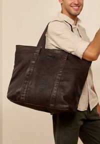 Dark brown leather tote bag with two long handles and stitched accents. Smooth texture with a rectangular shape. Subtle logo detail visible.
