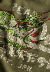 Camiseta oliva con un gráfico de dragón colorido decorado con brillantes cristales y texto audaz en colores contrastantes.