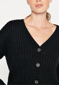 Svart stickad kofta med djup V-ringning, ribbat mönster och fem dekorativa knappar med metalliska detaljer.