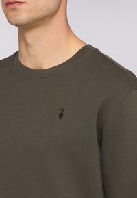 Olivgrön crew neck sweatshirt med ribbad textur och en diskret logotyp broderad på vänster bröst. Slät, mjuk material.