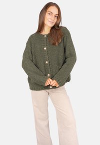 Grüne, flauschige Strickjacke mit strukturiertem Finish, ausgestattet mit großen Knöpfen und zwei frontalen Taschen, kombiniert mit hellen, weit geschnittenen Hosen.