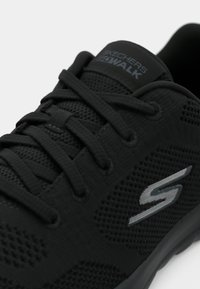 Zapato Skechers Go Walk negro con parte superior de malla, cordones planos, cuello acolchado y suela texturizada. Marca plateada en la lengüeta.