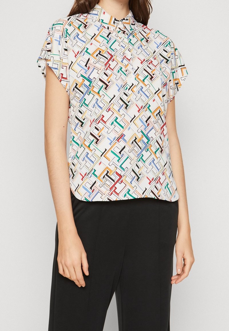 Chemise à manches courtes en tissu léger avec un motif géométrique coloré sur fond blanc, caractérisée par un col montant et une coupe décontractée.