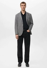 Szary, teksturowany blezer na czarnej koszulce polo, zestawiony z czarnymi spodniami i błyszczącymi butami. Blezer ma jedną kieszeń i zapinany jest na guzik.