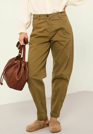 Olive green relaxed-fit pants with a button closure, side pockets, and rolled cuffs, paired with a brown leather bag and beige slip-on shoes.