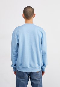 Sudadera azul con cuello y puños acanalados, que presenta un corte relajado y una textura suave. Combinada con jeans de mezclilla, con costuras visibles en los bolsillos.