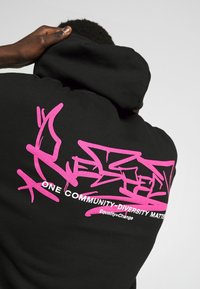 Svart huvtröja med en stor rosa graffiti-design på baksidan, med texten "ETT SAMHÄLLE-MANGFALD ÄR VIKTIGT."