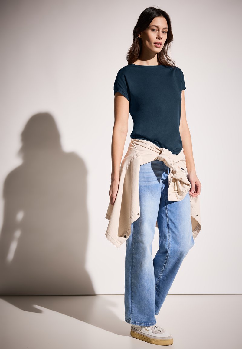 Dunkelbluse Kurzarmshirt kombiniert mit hellblauen weit geschnittenen Jeans, und einem hellbeigen Shirt, das um die Taille gebunden ist. Weiße Sneaker.