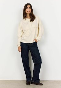 Maglietta crema a maniche lunghe con due tasche sul petto, abbottonatura e vestibilità comoda, abbinata a jeans a gamba larga blu scuro e scarpe marroni.
