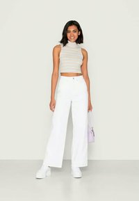 Jovem mulher vestindo um top cropped listrado sem mangas, calças largas brancas de cintura alta, tênis brancos e segurando uma pequena bolsa lavanda.