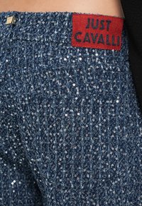 Blue sequin-embellished fabric with a textured pattern; features a red leather tag labeled "JUST CAVALLI" near the waistband.