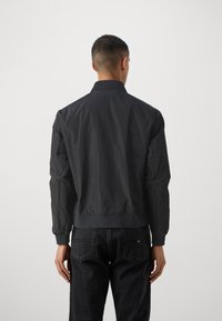 Veste bomber noire, en tissu léger, col et poignets côtelés, surface texturée, taille ajustée, associée à un pantalon en denim foncé, design minimaliste.
