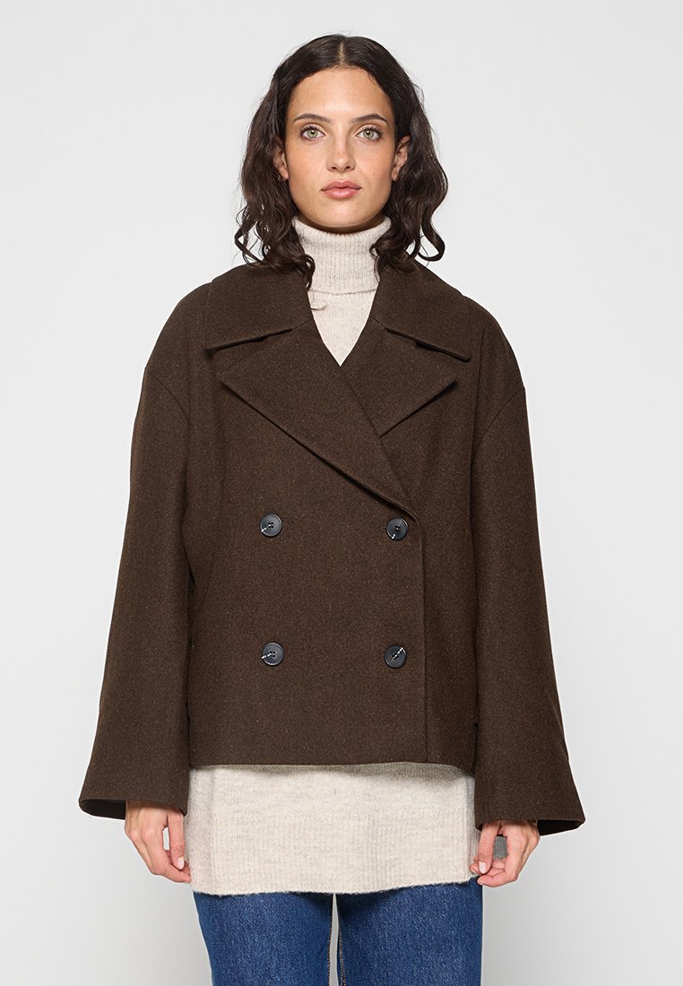 Cappotto marrone doppiopetto in lana, con ampi rever, bottoni neri e un design corto, abbinato a un dolcevita chiaro.