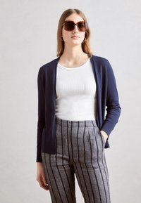 Un gilet marine sur une chemise blanche à côtes, associé à un pantalon rayé gris. Modèle portant de grandes lunettes de soleil brunes. Texture lisse, coupe décontractée.