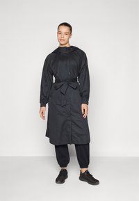 Long black windbreaker with a drawstring hood, cinched waist, and side pockets. Slim fit trousers and black athletic shoes complete the outfit.