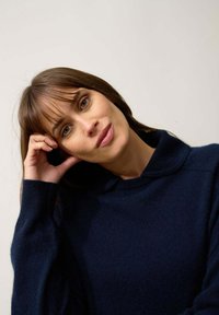 Granatowy sweter dziany z wysokim kołnierzem, miękką fakturą i długimi rękawami; modelka prezentująca swobodną pozę na neutralnym tle.