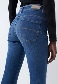Jeans in denim blu con un design aderente, caratterizzati da vita alta, tasche posteriori a toppa e cuciture arancioni sui bordi.