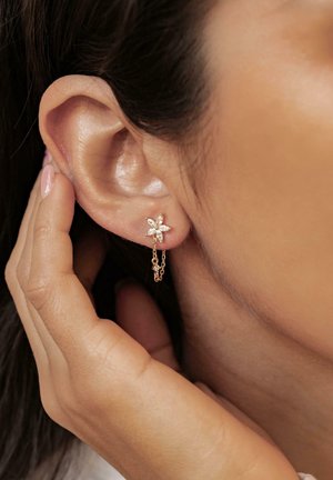 Golde Ohrringe mit floralem Design und markanten, klarsichtigen Steinen im Marquise-Schliff, die durch eine zarte Kette nahe am Ohr befestigt sind. Glatte metallische Oberfläche.