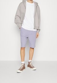 Personne portant un sweat à capuche gris à fermeture éclair, un t-shirt blanc, un short violet clair, des chaussettes blanches et des baskets montantes marron, debout sur un fond uni.