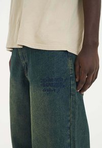 Béžové bavlněné tričko v kombinaci s tmavě zelenými džíny s texturovaným vyšitým logem na levém stehně.