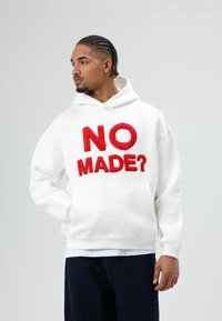 Witte hoodie met grote rode tekst "NO MADE?" op de voorkant. Voorzien van een kangoeroezak en een capuchon met trekkoord. Model draagt donkere spijkerbroek.