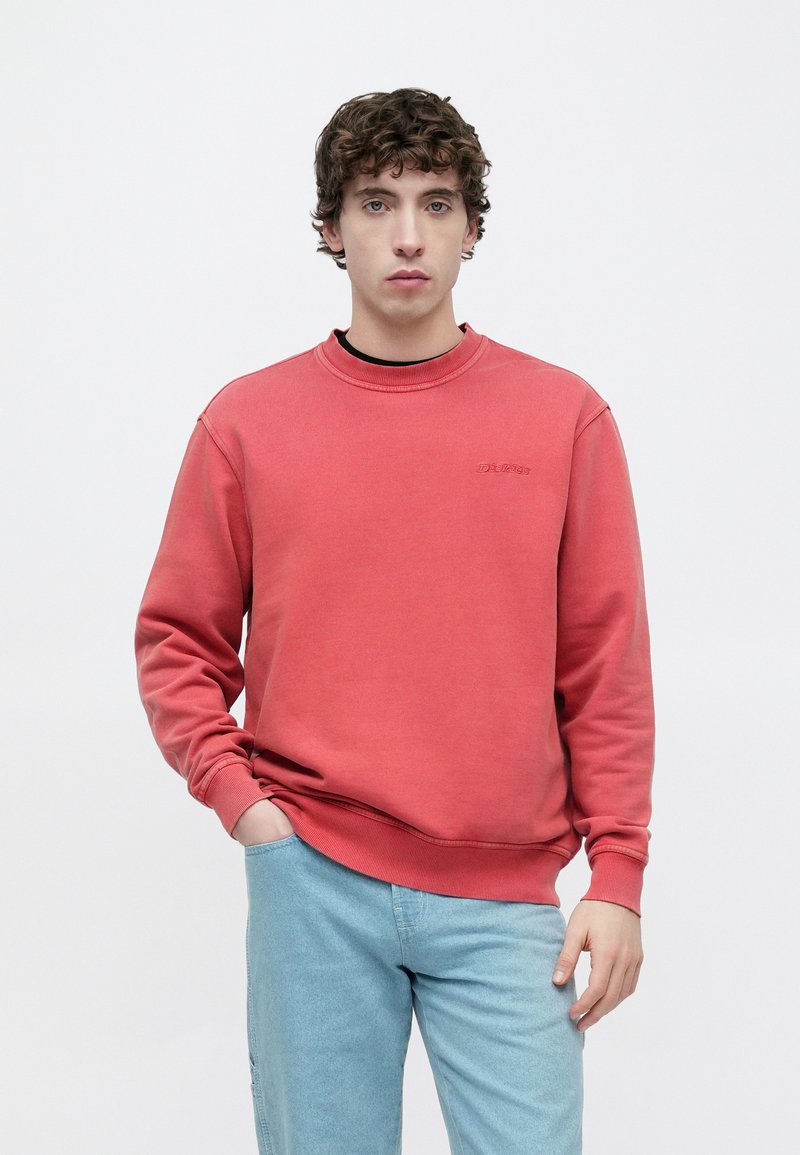 Felpa rossa in cotone con scollo tondo e polsini a costine, con un piccolo logo del marchio ricamato sul petto. Indossata con jeans azzurri.