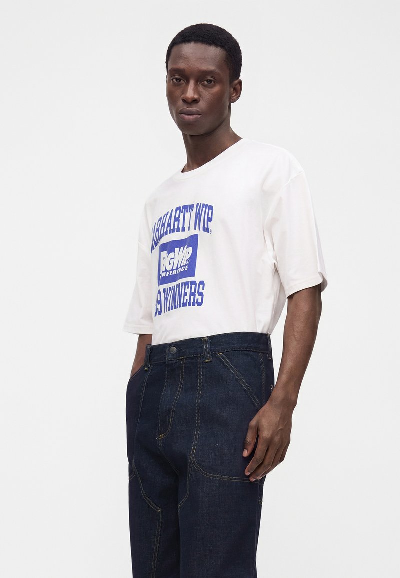 Man wearing white short-sleeve shirt with blue lettering and dark blue jeans, standing with one hand in pocket against plain background.