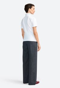Top corto bianco a maniche corte con colletto arricciato, abbinato a pantaloni a gamba larga grigio scuro. Modella di spalle, in piedi su uno sfondo neutro.
