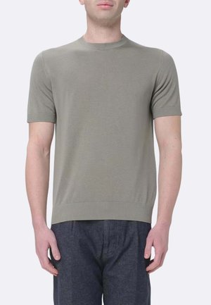 Uomo che indossa una maglietta a maniche corte in maglia verde oliva chiaro e pantaloni blu scuro, in piedi davanti a uno sfondo chiaro e semplice.