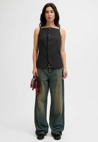 Kvinna med långt mörkt hår, iförd svart ärmlös knäppt topp, vida bleka jeans, svarta skor och håller en liten vinröd handväska.