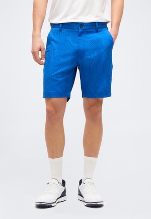 Άνδρας με μπλε σορτς με σχέδια, λευκές κάλτσες και αθλητικά παπούτσια λευκά και μαύρα, στέκεται με τα χέρια στις τσέπες.